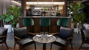 A bar area with a counter, bar stools, and a variety of bottles. There are two chairs and a table in the foreground. - Servotel Saint-Vincent (Nice)