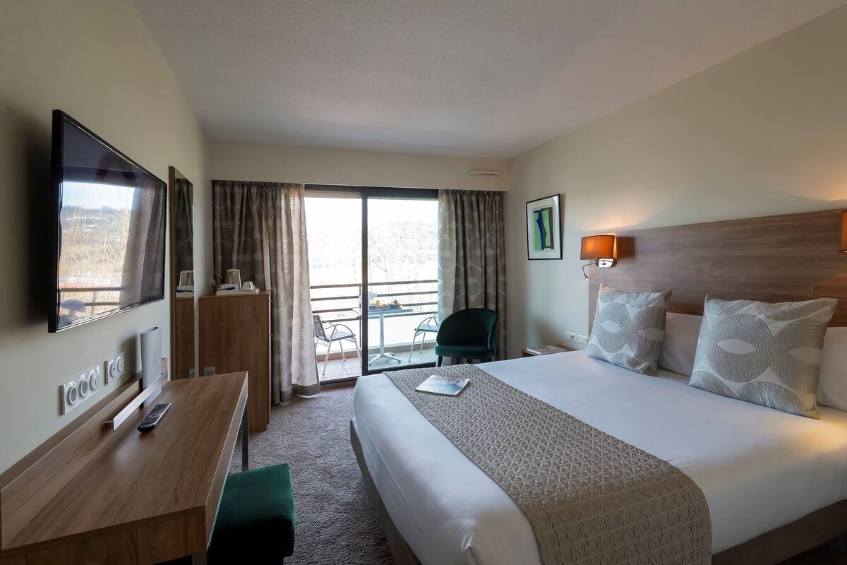 A hotel room with a large bed, a desk with a TV, a chair, and a balcony with a view.