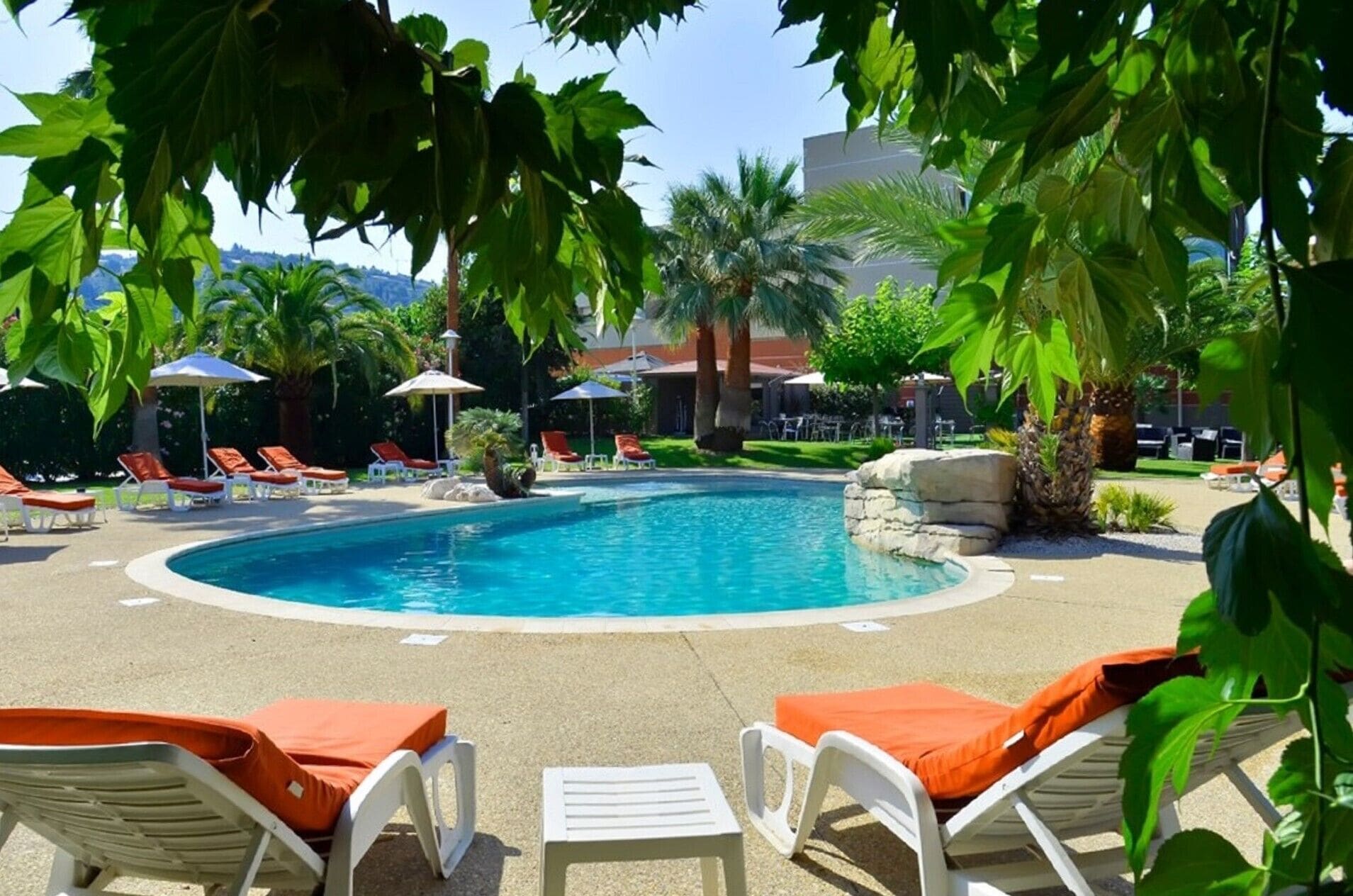 A swimming pool with lounge chairs and umbrellas, surrounded by greenery and palm trees.