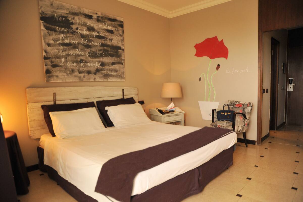 Een hotelkamer met een groot bed, een nachtkastje met een lamp, een stoel en een muur met een schilderij van een rode bloem.