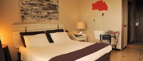 A hotel room with a large bed, a bedside table with a lamp, a chair, and a wall with a red flower painting.