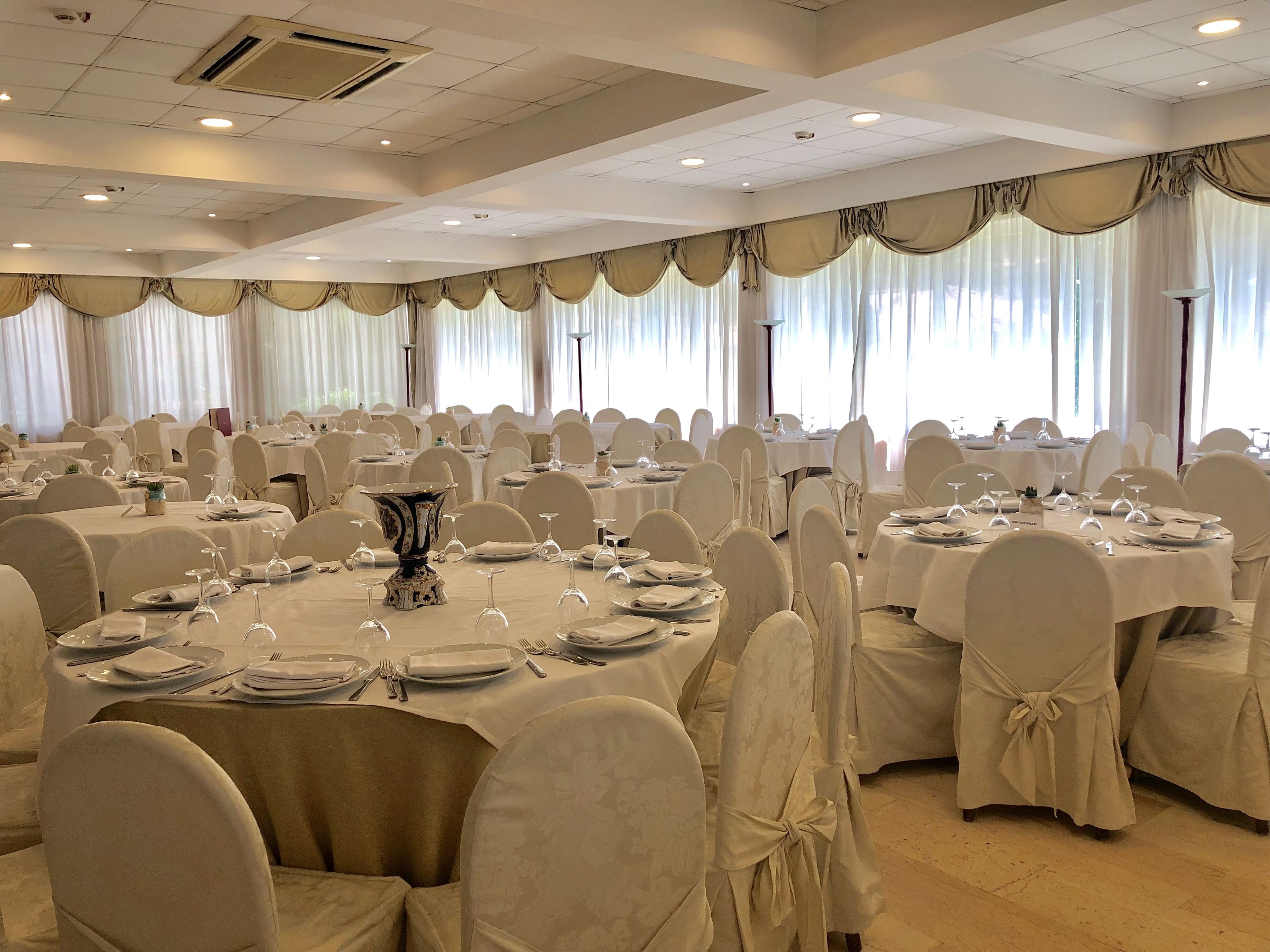 Une salle de banquet avec des tables rondes dressées pour le repas, des chaises blanches et de grandes fenêtres ornées de rideaux.