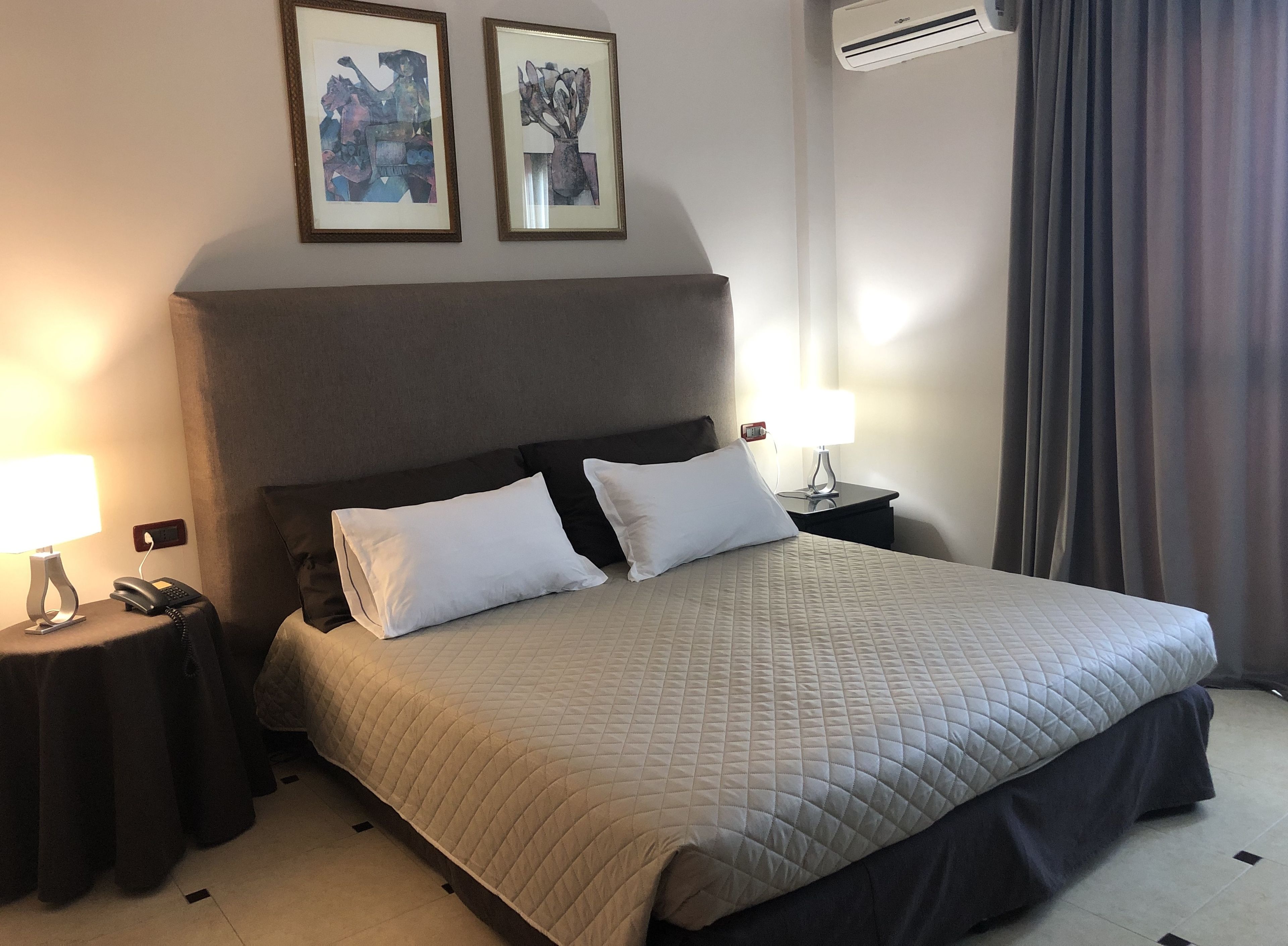 A hotel room with a large bed, two bedside tables with lamps, a phone, and two framed artworks on the wall.