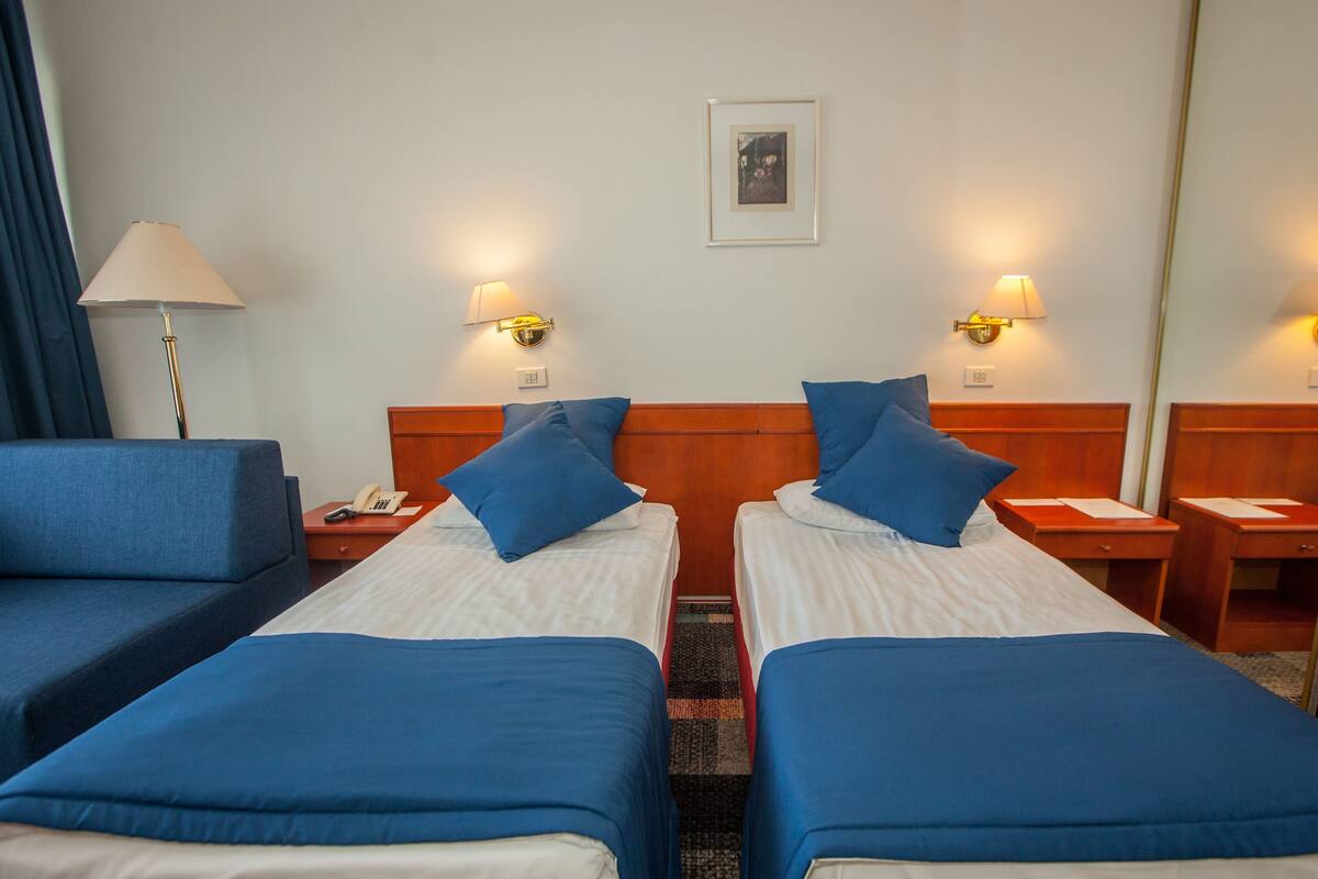 A hotel room with two single beds, a sofa, a nightstand with a lamp, and a framed picture on the wall.