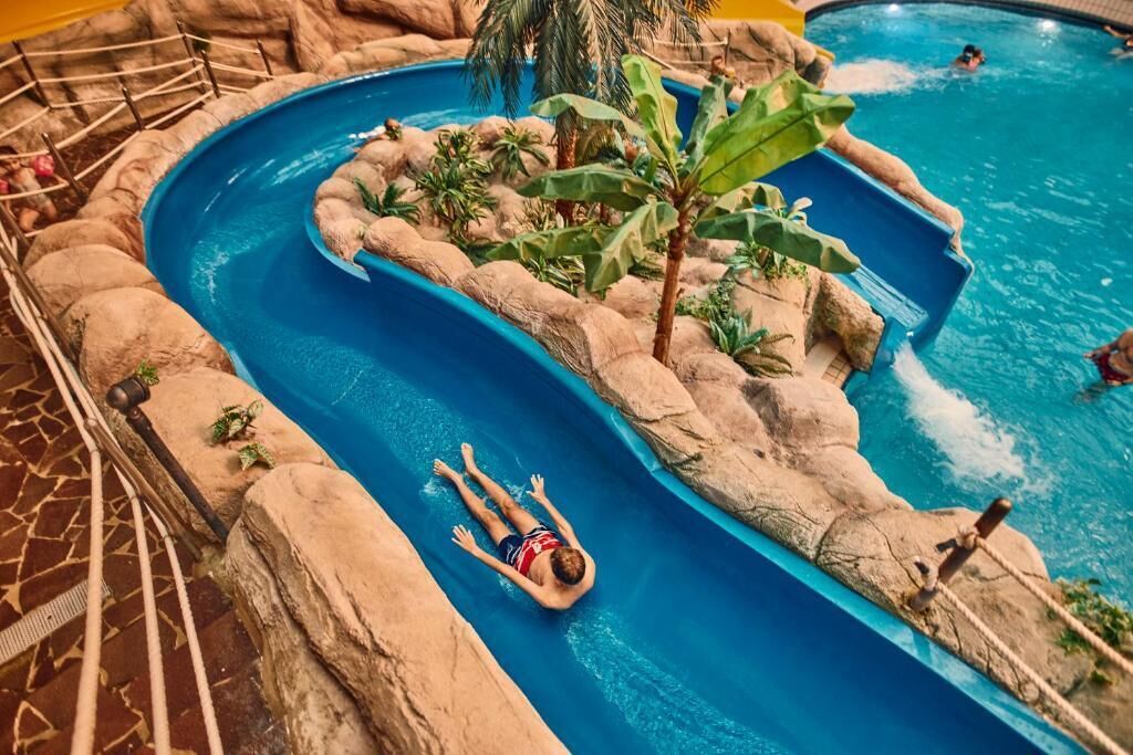 Piscina interna com escorregador, rodeada por pedras e plantas artificiais.