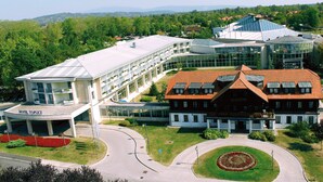 Un hotel con fachada de vidrio moderno, un edificio tradicional de techo rojo y un jardín ajardinado con un parterre circular.