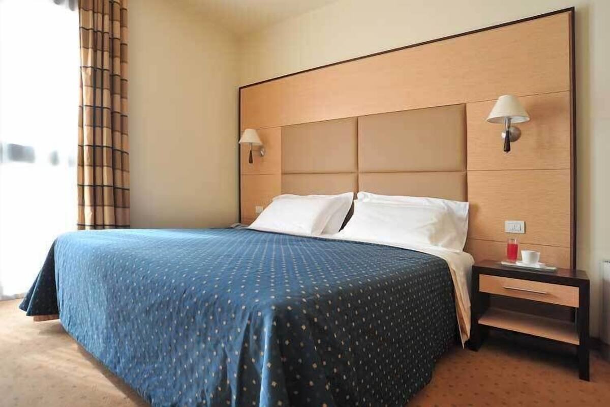 A hotel room with a large bed, two bedside lamps, a nightstand with a cup and saucer, and a window with curtains.