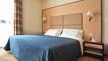 Una habitación de hotel con una cama grande, dos lámparas de noche, una mesita de noche con una taza y un platillo, y una ventana con cortinas.