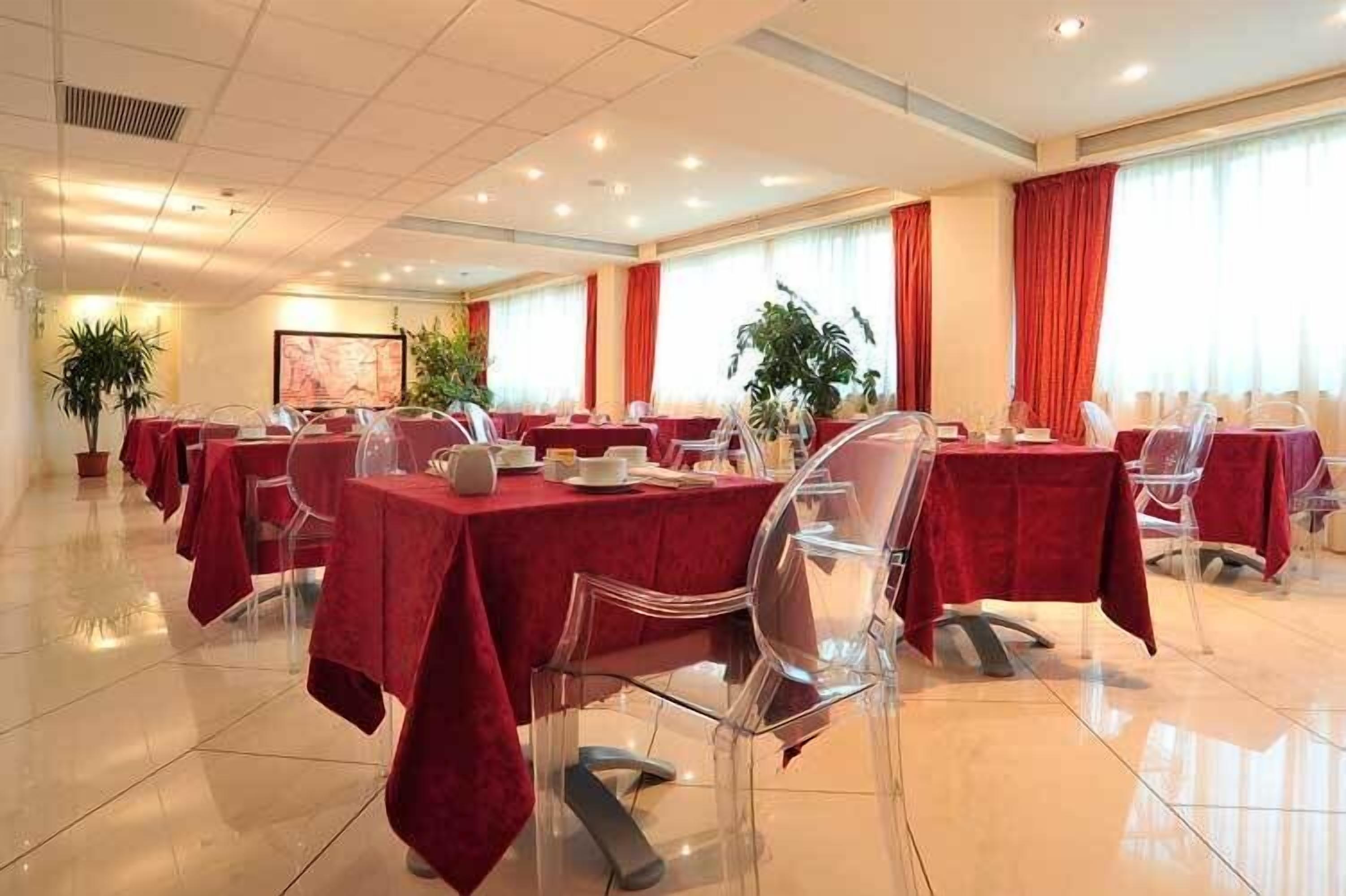 Un salón de conferencias con mesas cubiertas de manteles rojos, sillas traslúcidas y plantas en macetas.
