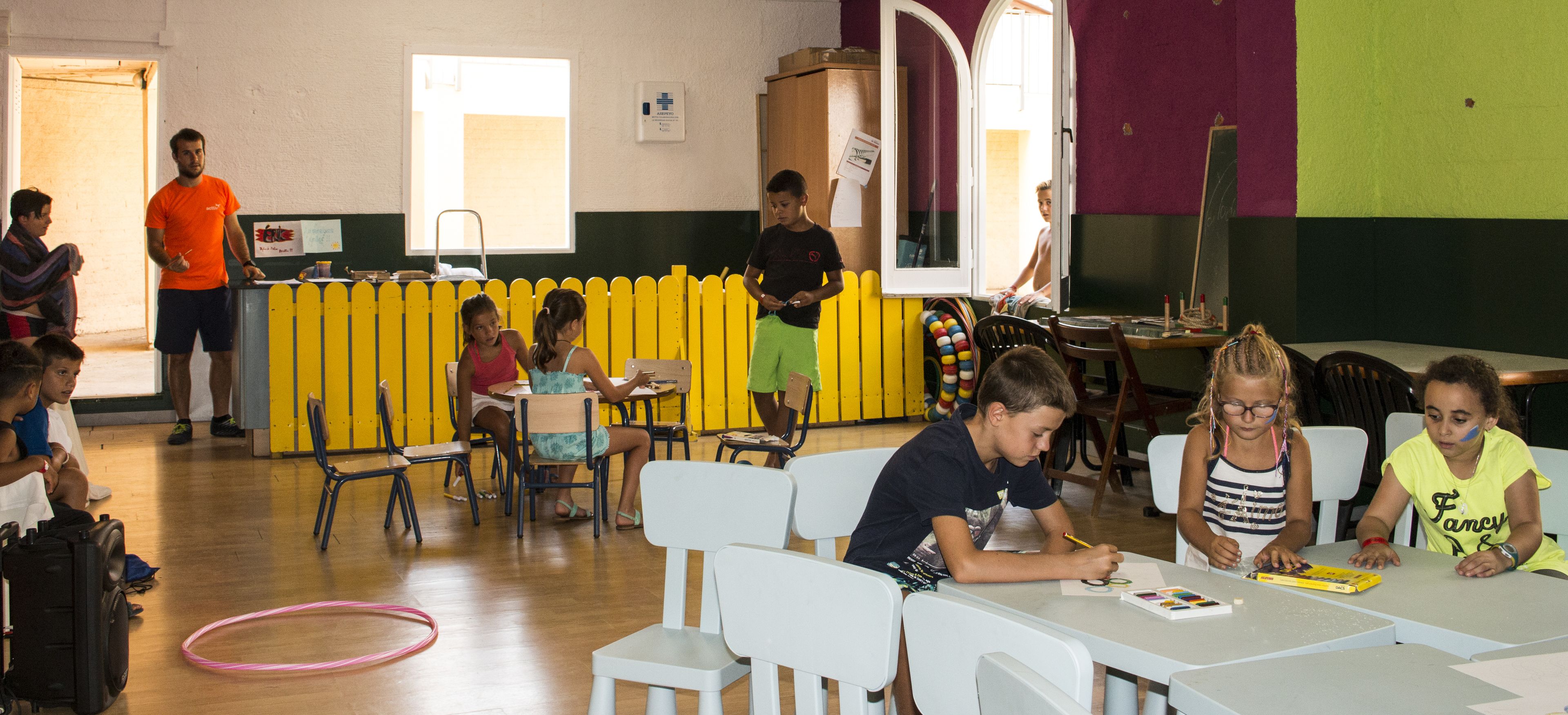 Une salle de classe où des enfants sont assis à des tables, un enseignant se tient près d’un tableau noir, et une clôture jaune est visible à l’arrière-plan.