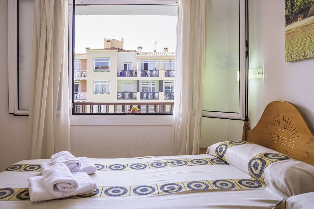 A hotel room with a bed, a window with curtains, and a view of a building with balconies.