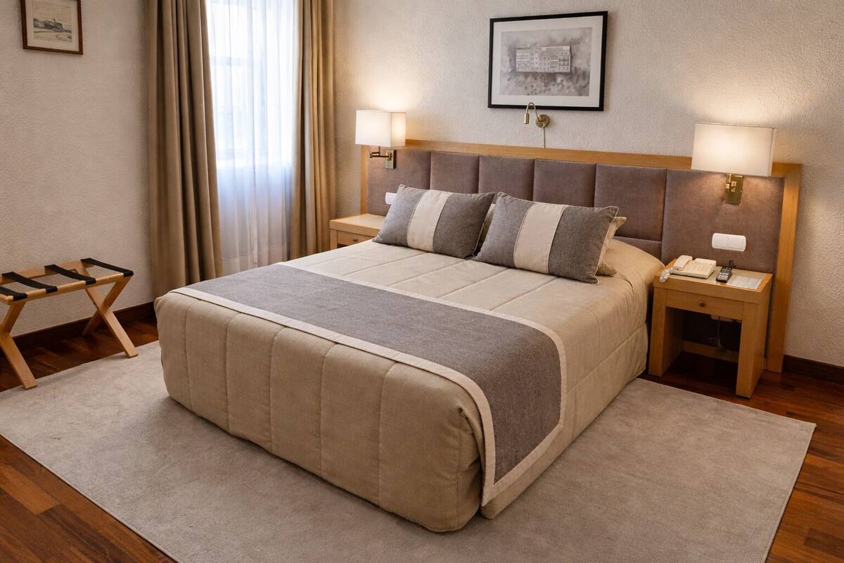 A neatly arranged hotel room with a large bed, two bedside tables, a wooden nightstand, a small table, and a window with curtains.