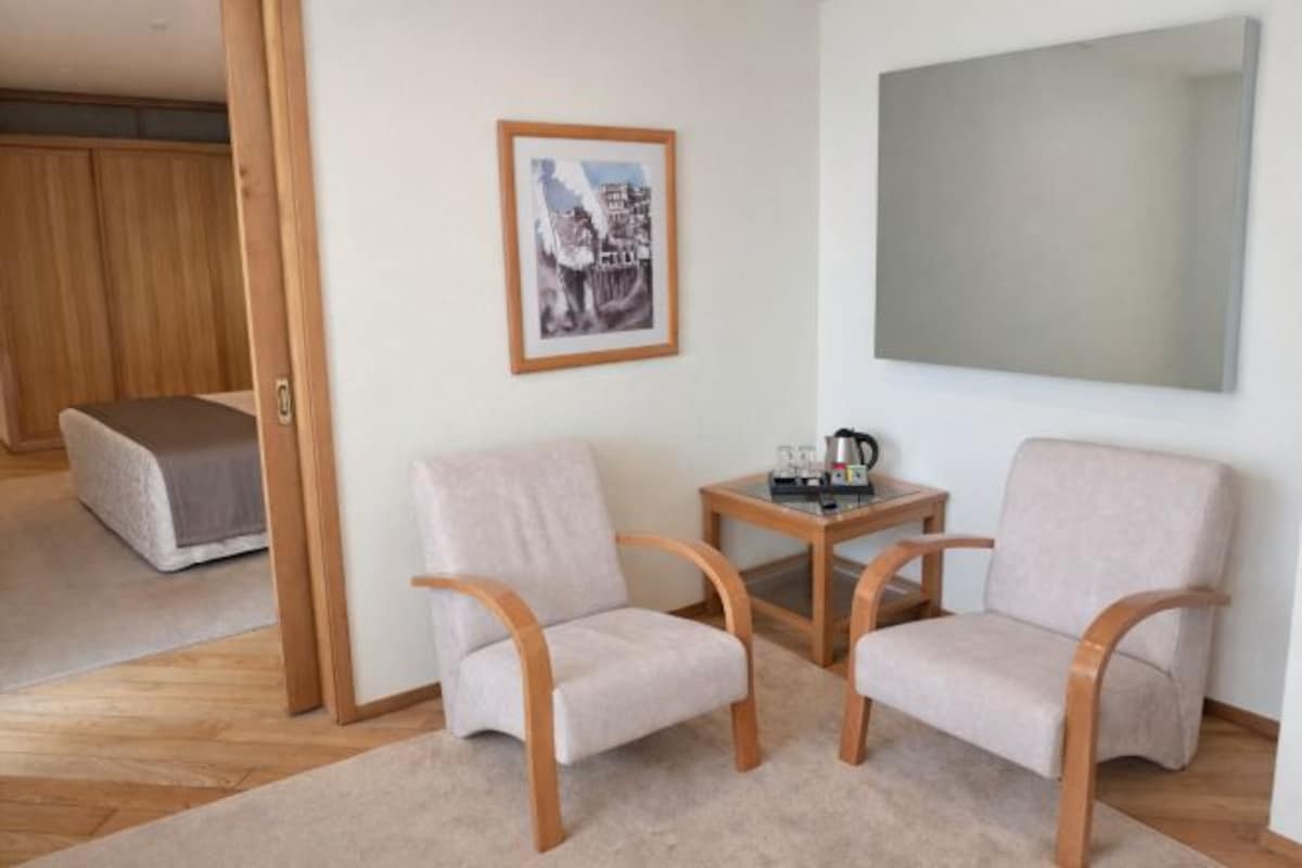 A room with two light-colored armchairs, a small wooden table with a coffee pot, and a framed picture on the wall.
