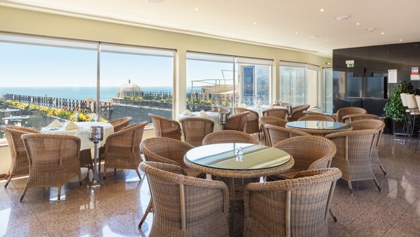 A restaurant with wicker chairs and tables, large windows with a view of the sea, and a modern interior design. - Hotel Boa-Vista (Porto)