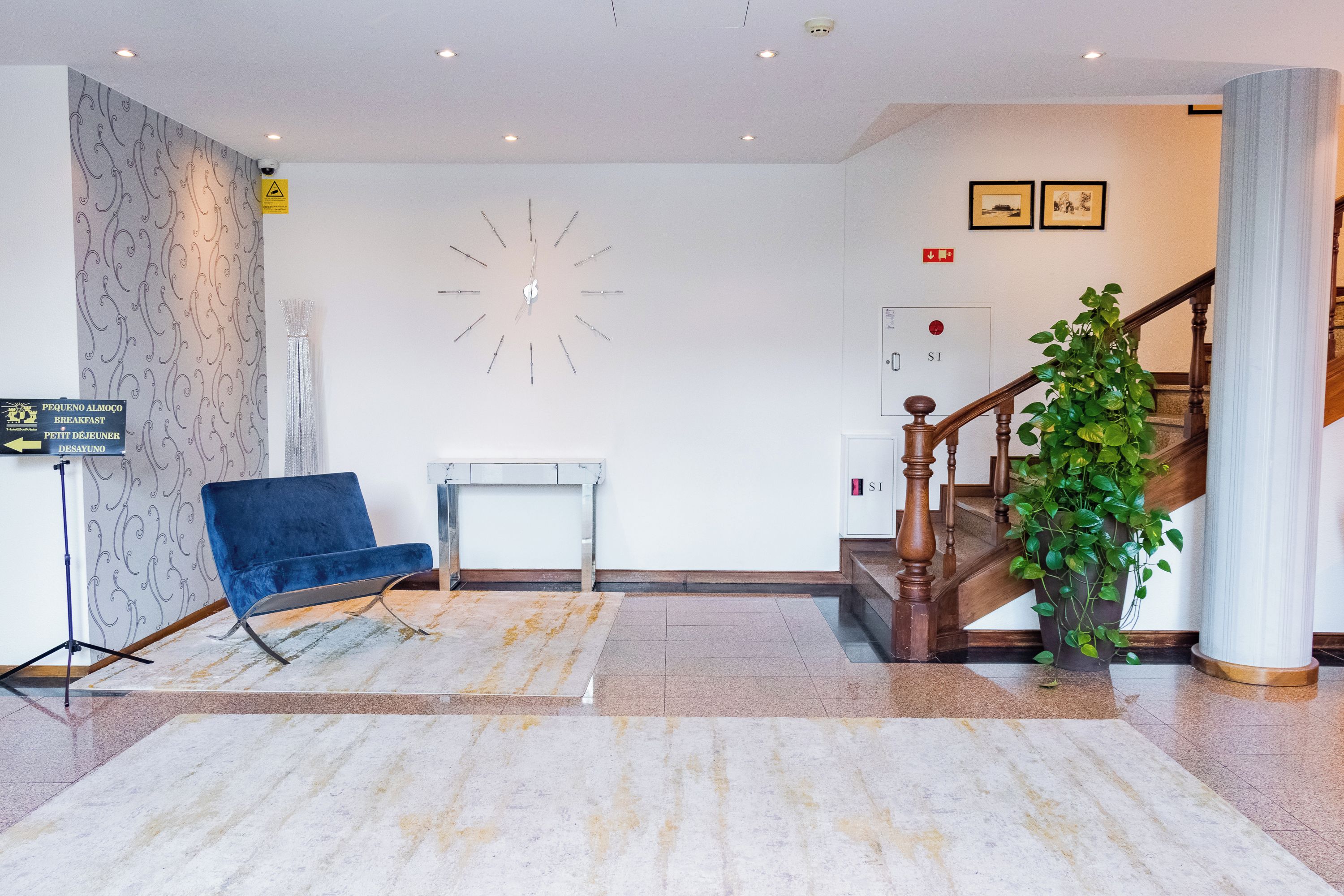 Un hall d’hôtel avec un fauteuil bleu, une horloge, un escalier et une plante.