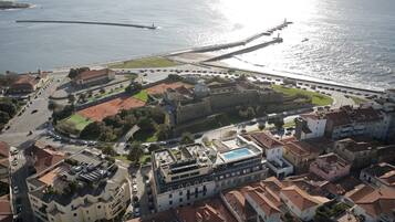 Vista aérea de um hotel à beira-mar com piscina, uma construção fortificada e uma estrada que leva à praia.