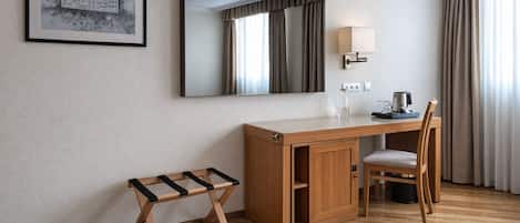 A hotel room with a wooden desk, a chair, a small stool, a mirror, a lamp, and a framed picture on the wall.