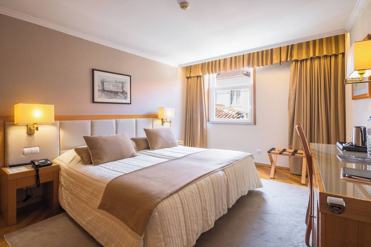 A hotel room with a large bed, bedside lamps, a desk with a chair, a window with curtains, and a framed picture on the wall.