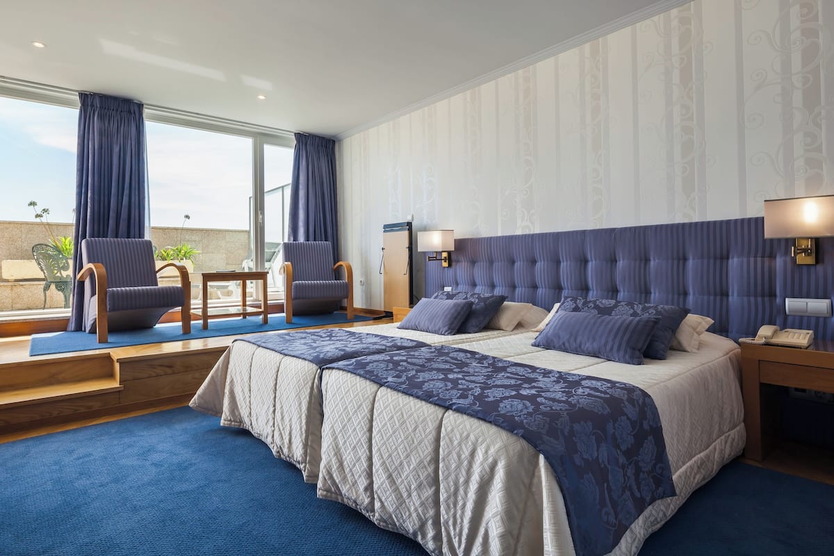 A hotel room with a large bed, two armchairs, a small table, and a view of the outdoors.