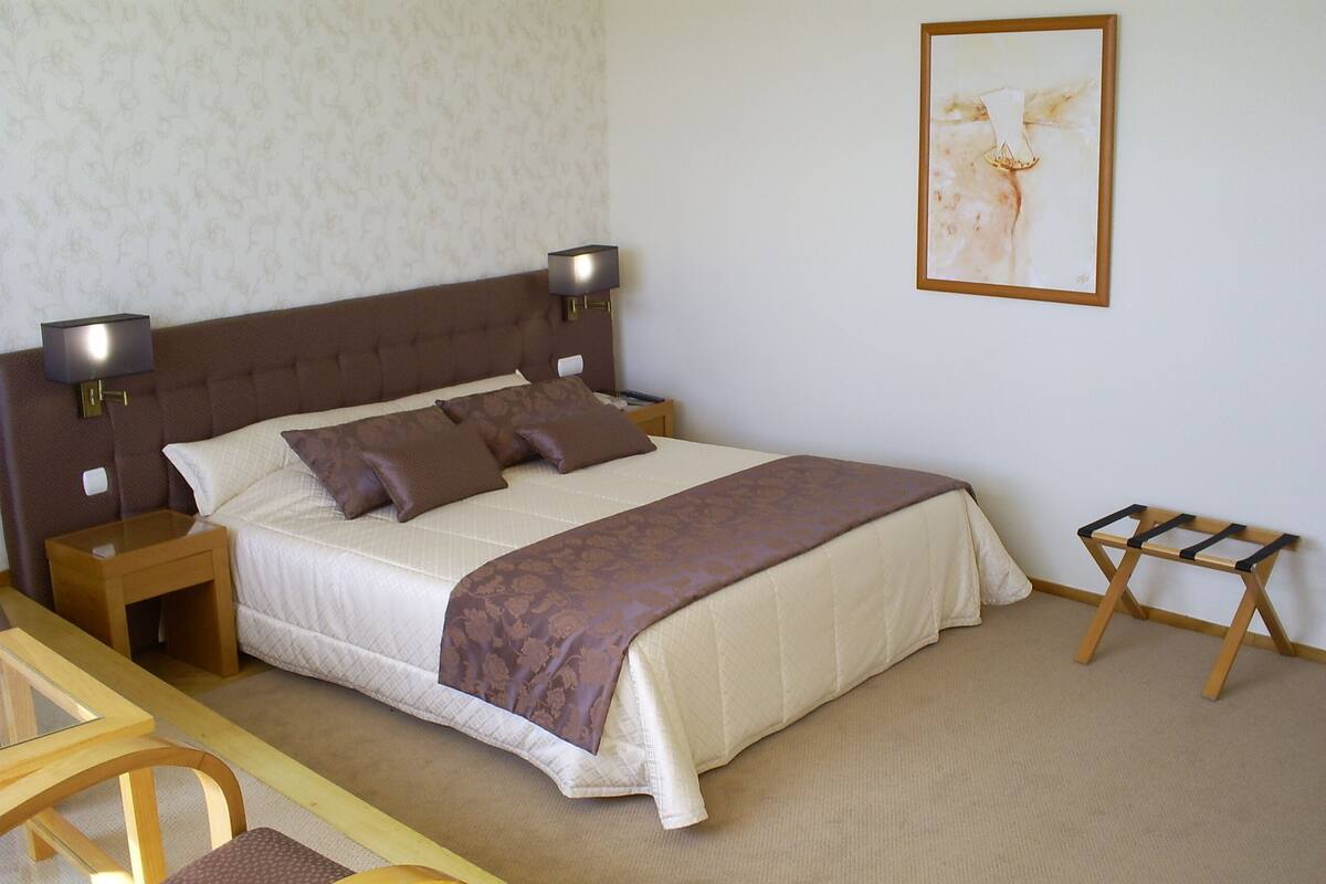 A neatly arranged bedroom with a bed, bedside tables, a small wooden stool, and a framed artwork on the wall.