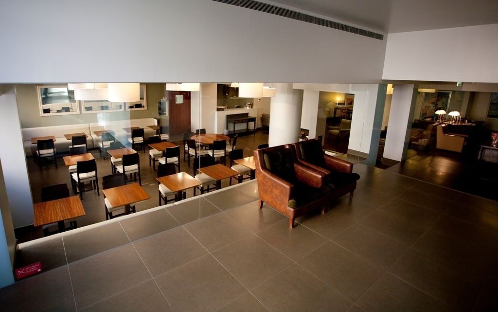 A spacious hotel lobby with multiple seating areas, including tables and sofas, and a visible kitchen area in the background.