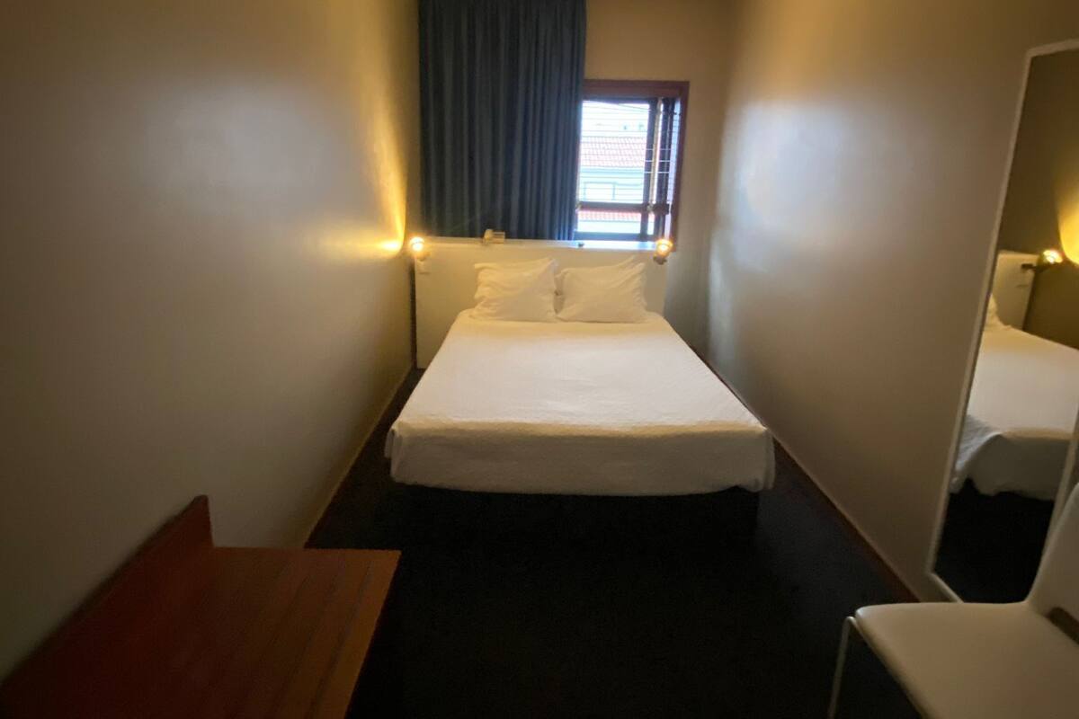 Una habitación de hotel pequeña y tenuemente iluminada, con una cama individual, una mesa de madera y un espejo.