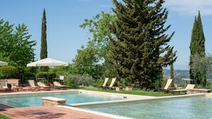 A rectangular swimming pool with lounge chairs and umbrellas, surrounded by tall trees and a clear sky. - Fonte de' Medici (San Casciano in Val di Pesa)