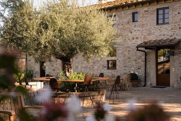 Un área de comedor al aire libre con muros de piedra y un árbol grande, con mesas y sillas.