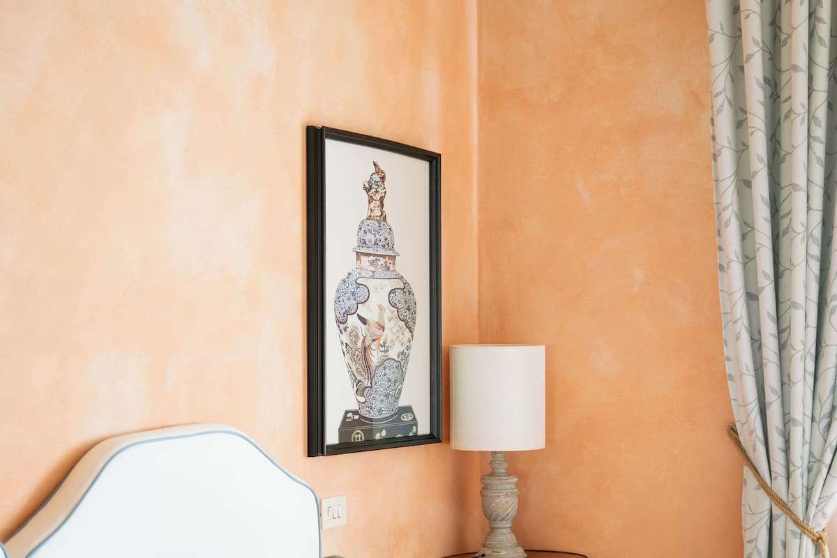 Une chambre à coucher avec un lit, des oreillers, une table de chevet avec une lampe, une œuvre d’art encadrée au mur et un rideau.