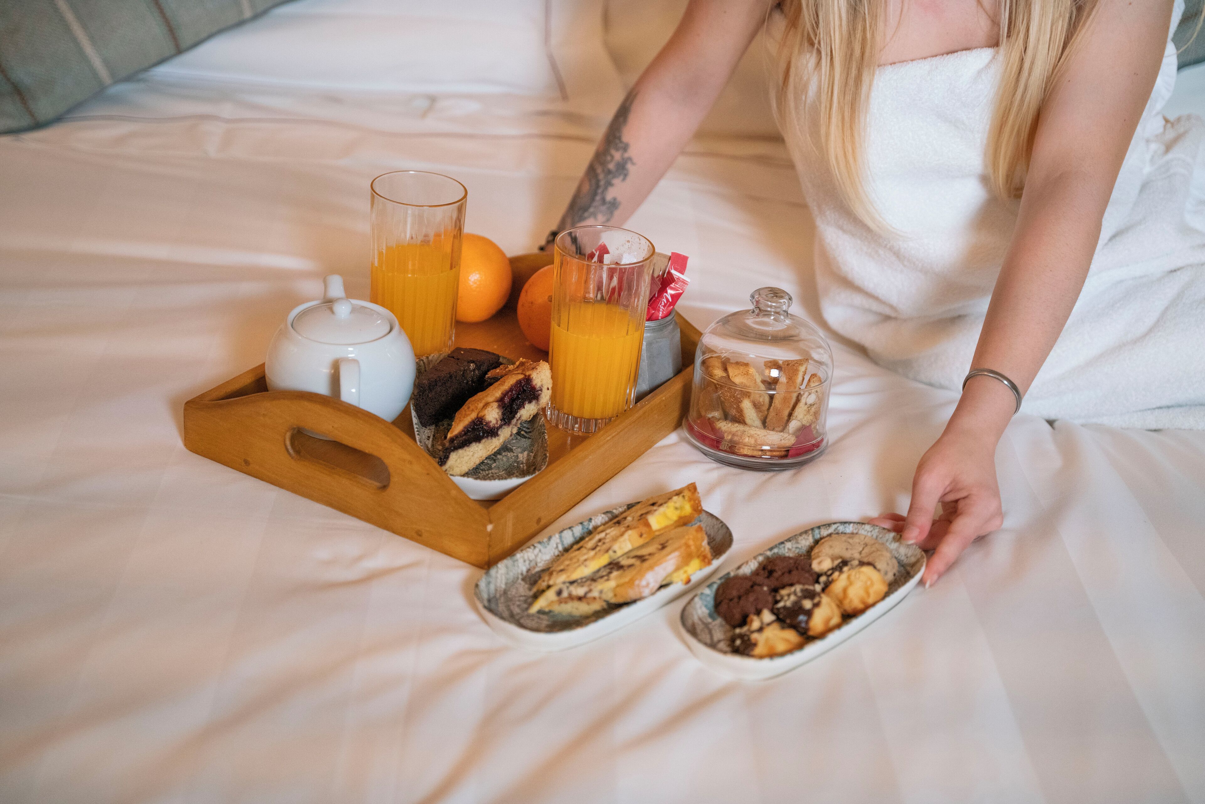 A breakfast tray with pastries, a teapot, orange juice, and a glass of orange juice on a bed.