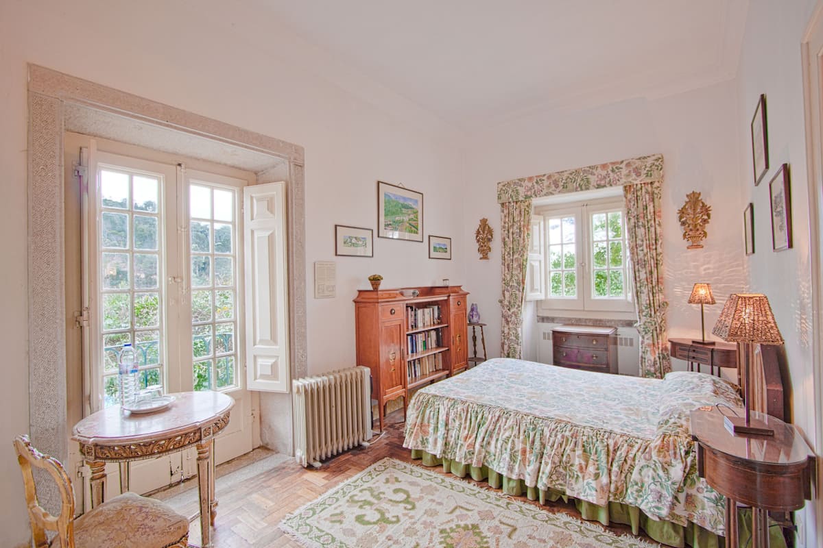 Een slaapkamer met een bed, een radiator, een houten kast, een tafel met stoel en een raam met gordijnen.
