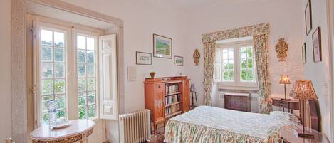 A bedroom with a bed, a radiator, a wooden cabinet, a table with a chair, and a window with curtains.