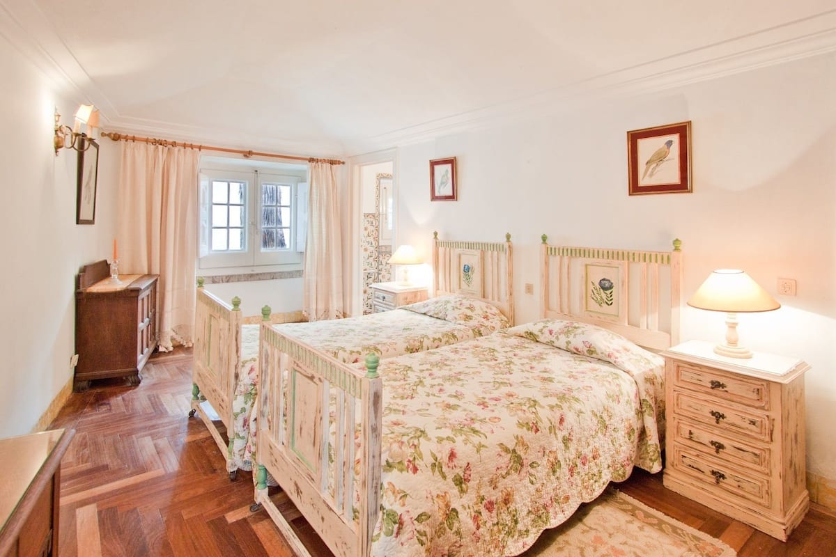 Een slaapkamer met twee bedden, een kaptafel, nachtkastje, lamp, raam met gordijnen en een muur met ingelijste foto’s.