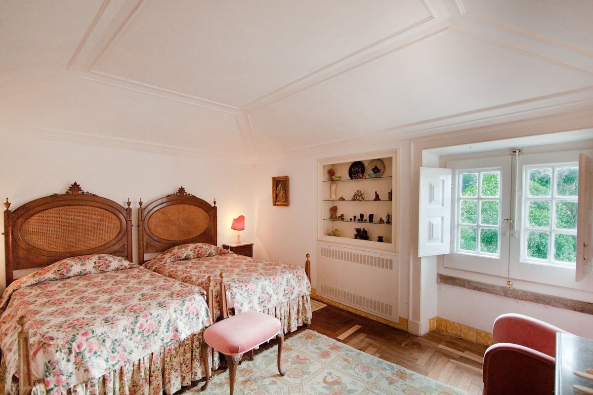 Slaapkamer met twee houten bedden, een klein tafeltje, een stoel, een raam met open luiken en een plank met decoratieve objecten.