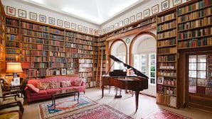 A spacious library with high ceilings, wooden bookshelves filled with books, a grand piano, a red sofa, and ornate rugs.