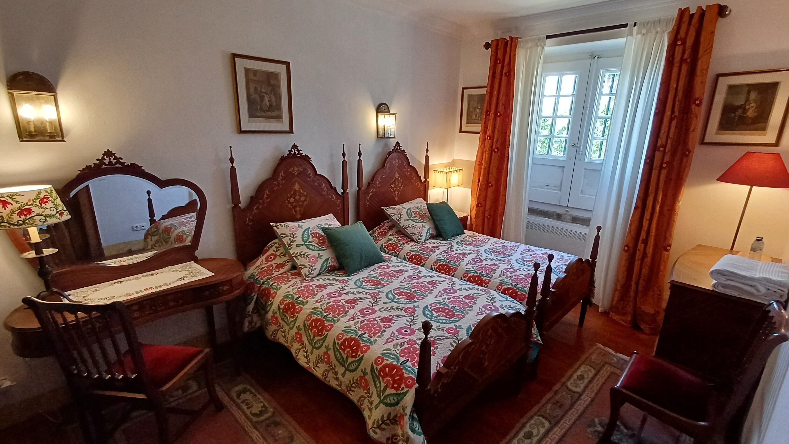 Un dormitorio tradicional con dos camas de madera, un escritorio y una silla. Hay lámparas fijadas a la pared y una puerta con cortinas.