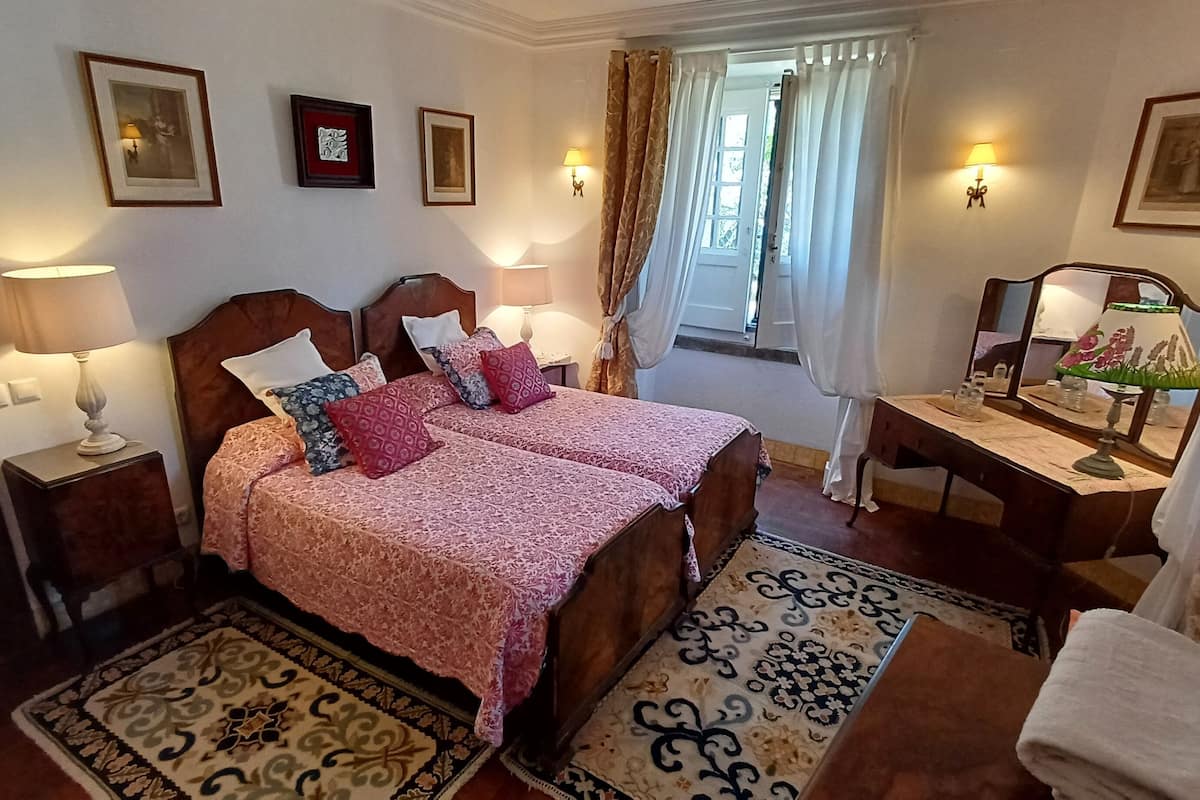 Een tweepersoonsbed met beddengoed met een patroon, twee nachtkastjes met lampen, een bureau met spiegel, en een raam met gordijnen.
