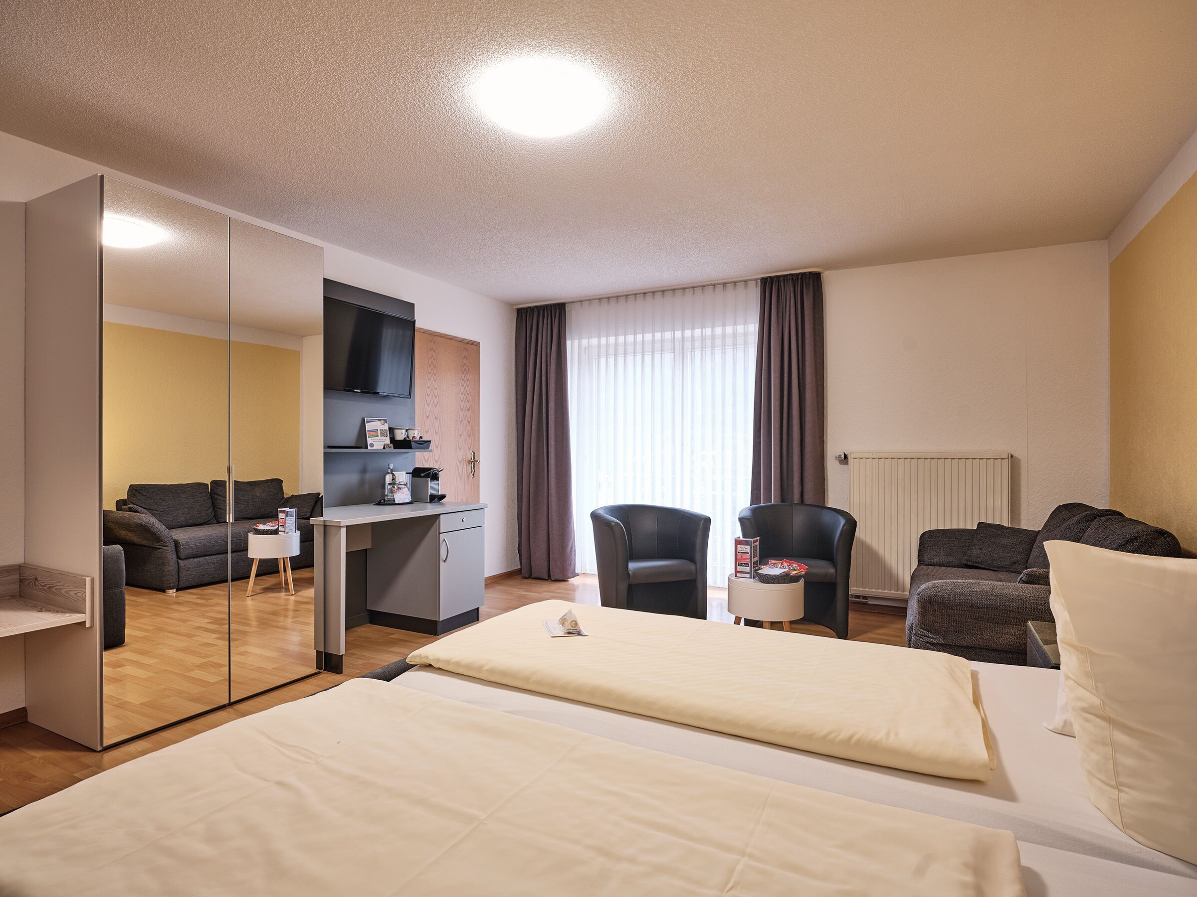 A hotel room with a bed, two armchairs, a desk with a flat-screen TV, a coffee machine, and a mirror.