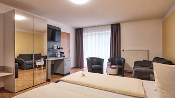 A hotel room with a bed, two armchairs, a desk with a flat-screen TV, a coffee machine, and a mirror.