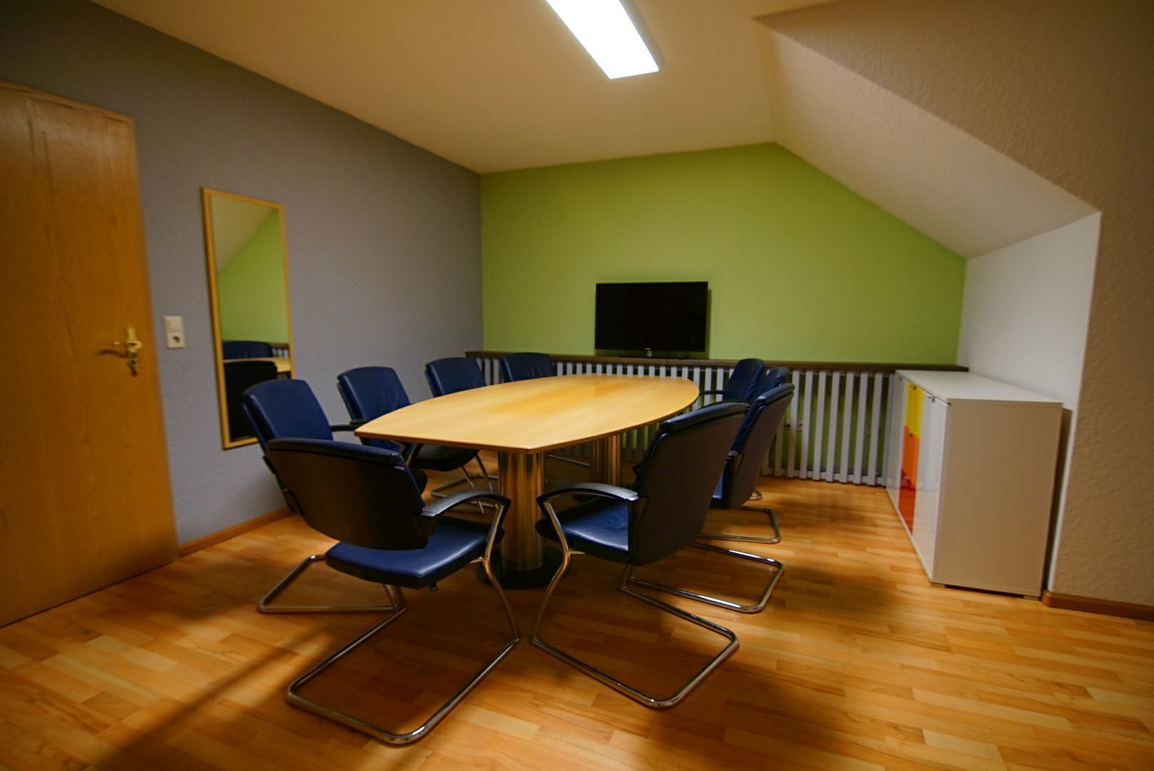 Une salle de conférence avec une table en bois et des chaises bleues, un téléviseur fixé au mur et un coin cuisine.