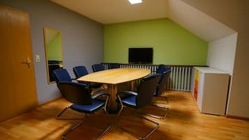 Une salle de conférence avec une table en bois et des chaises bleues, un téléviseur fixé au mur et un coin cuisine.