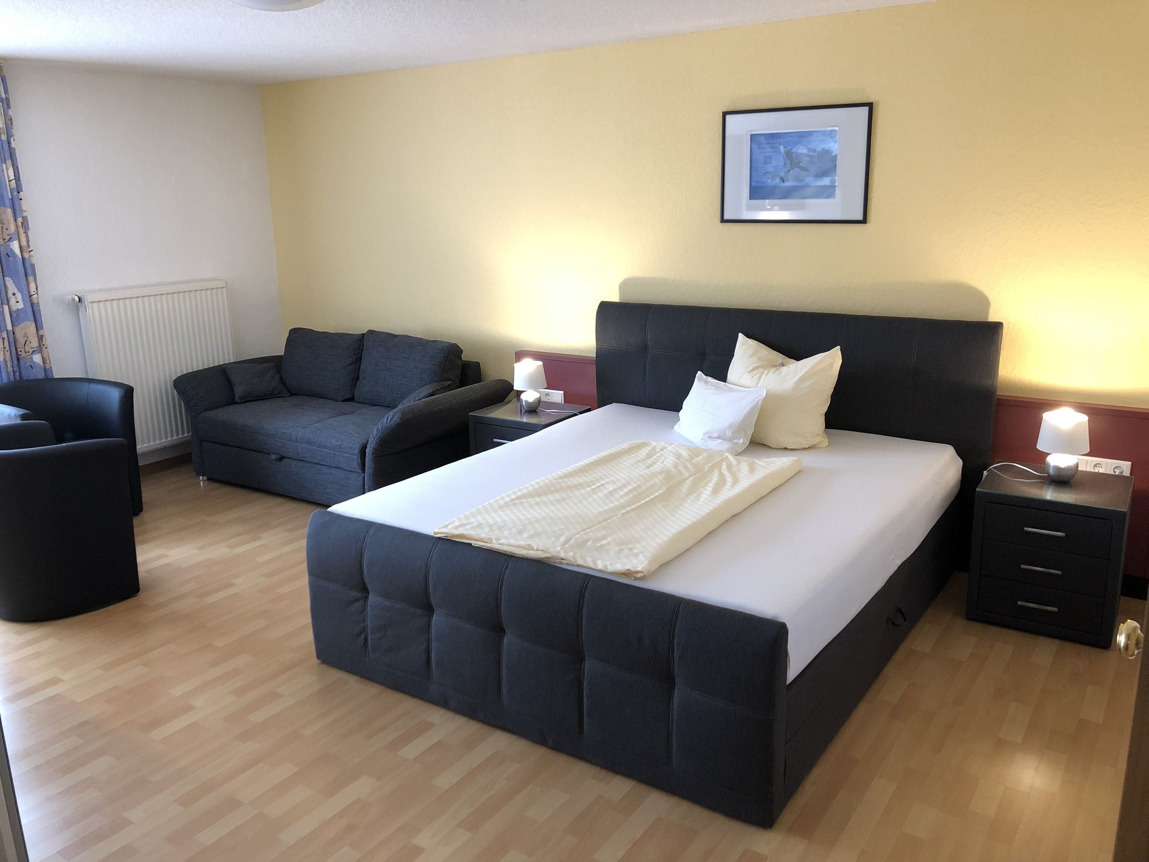 A hotel room with a large bed, a sofa, a chair, and a bedside table with a lamp.