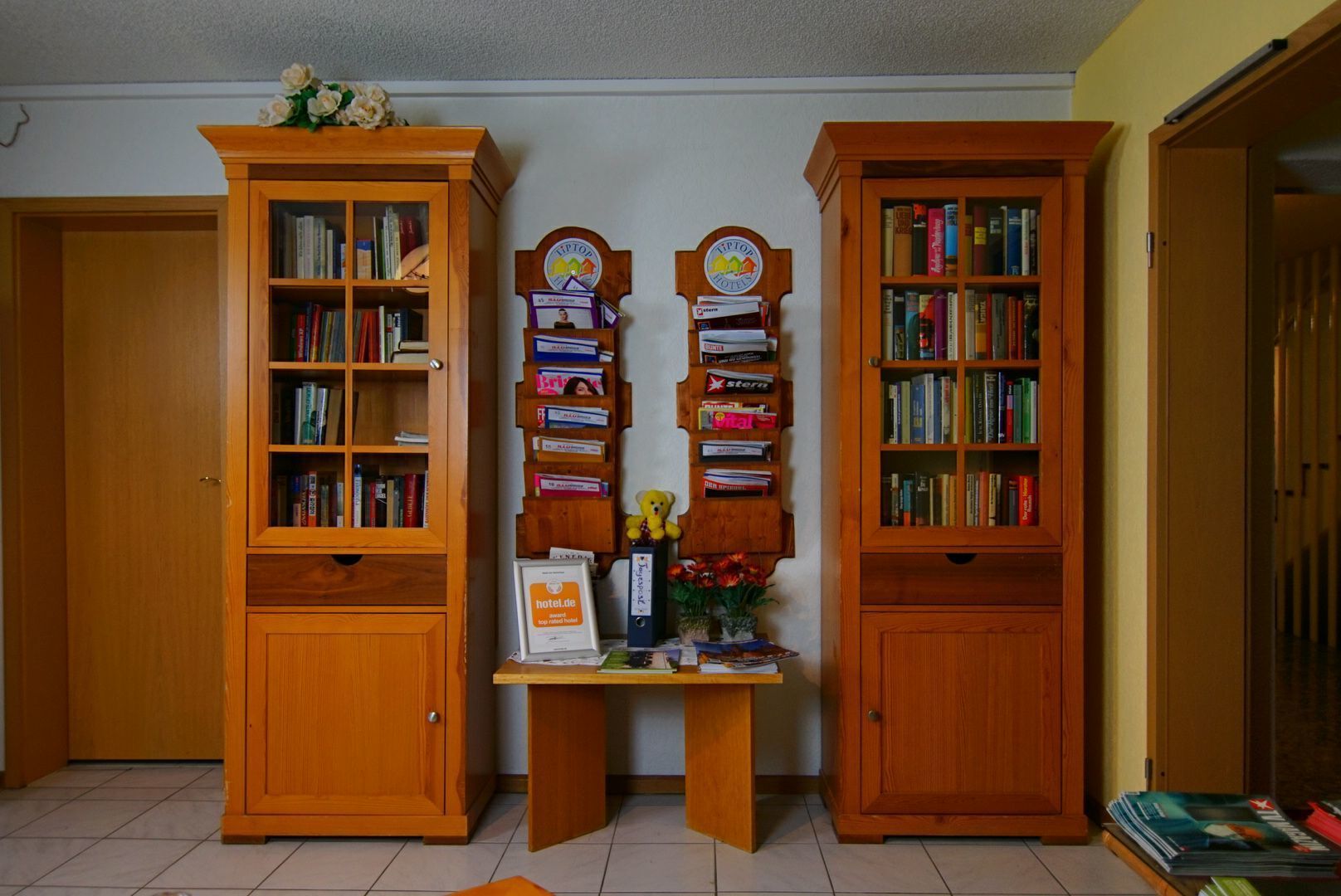 Une pièce avec deux étagères en bois remplies de livres, une petite table sur laquelle est posé un vase de fleurs, et une enseigne indiquant « hotel.de ».