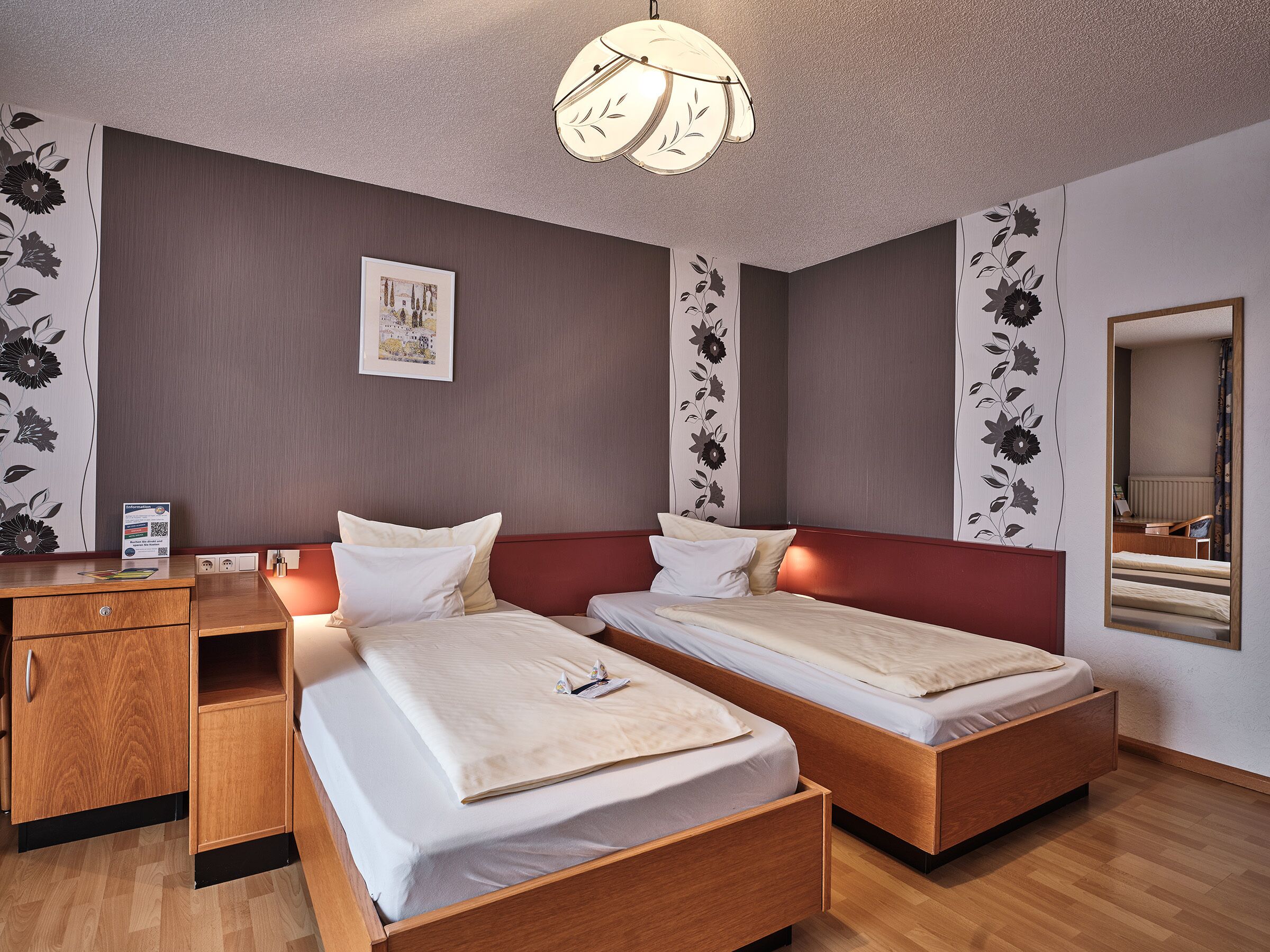 A hotel room with two single beds, a wooden desk, a mirror, and floral wall decorations.