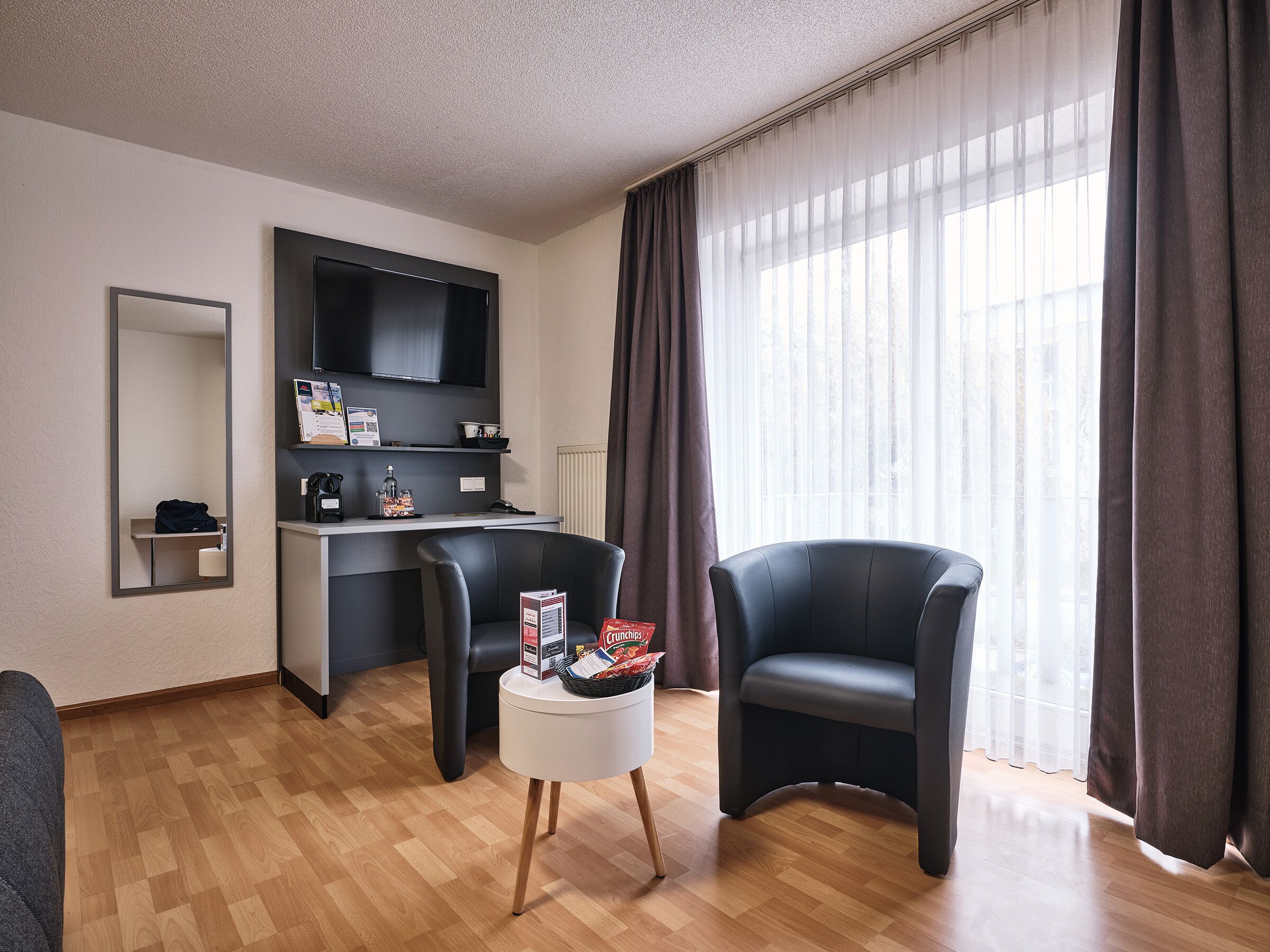 A modern living room with a flat-screen TV, a small kitchen area, two black armchairs, a white side table with snacks, and a large window with curtains.