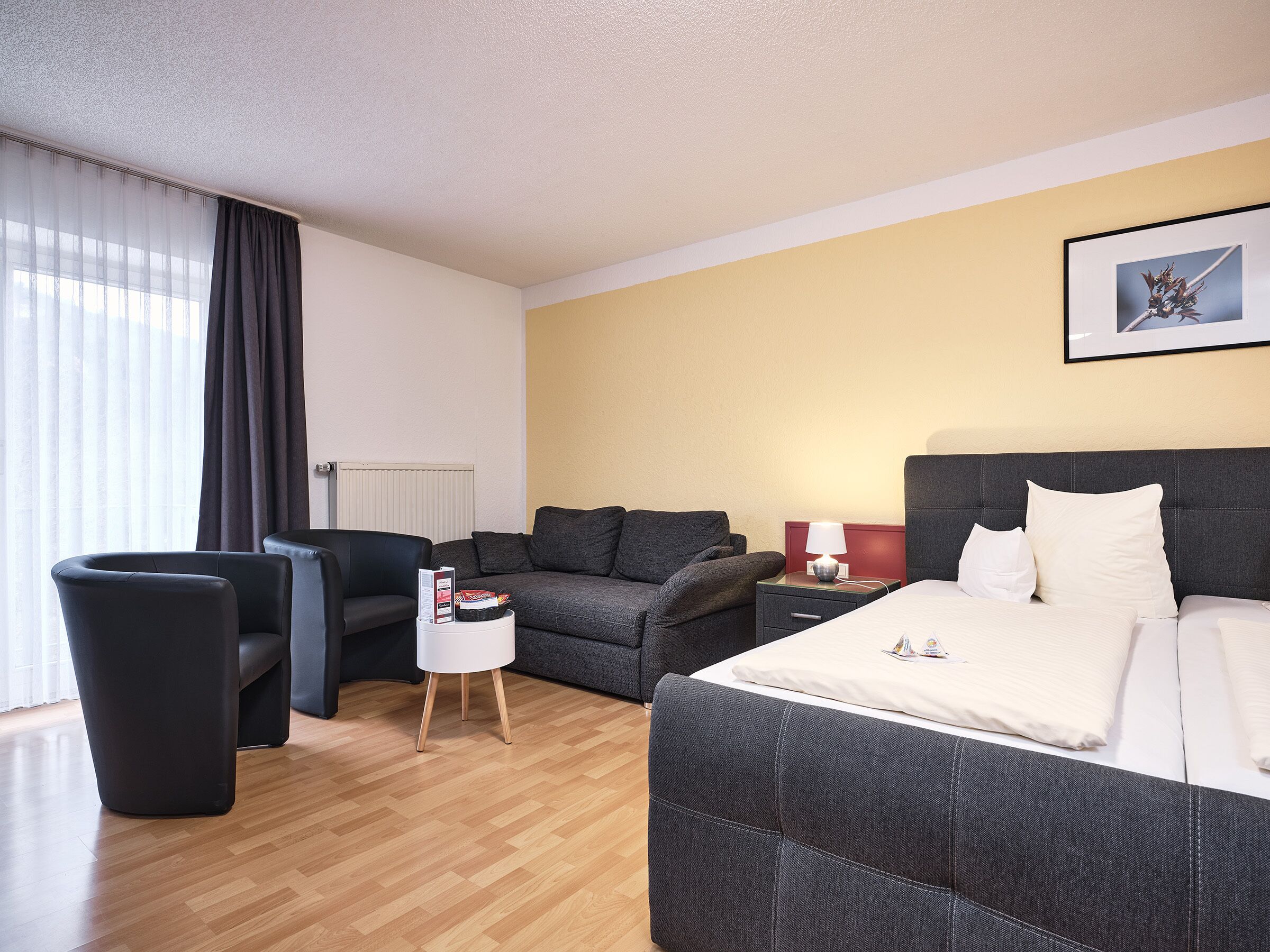 Ein Hotelzimmer mit einem Bett, einem Sofa, einem Sessel, einem Nachttisch mit Lampe sowie einem gerahmten Bild an der Wand.