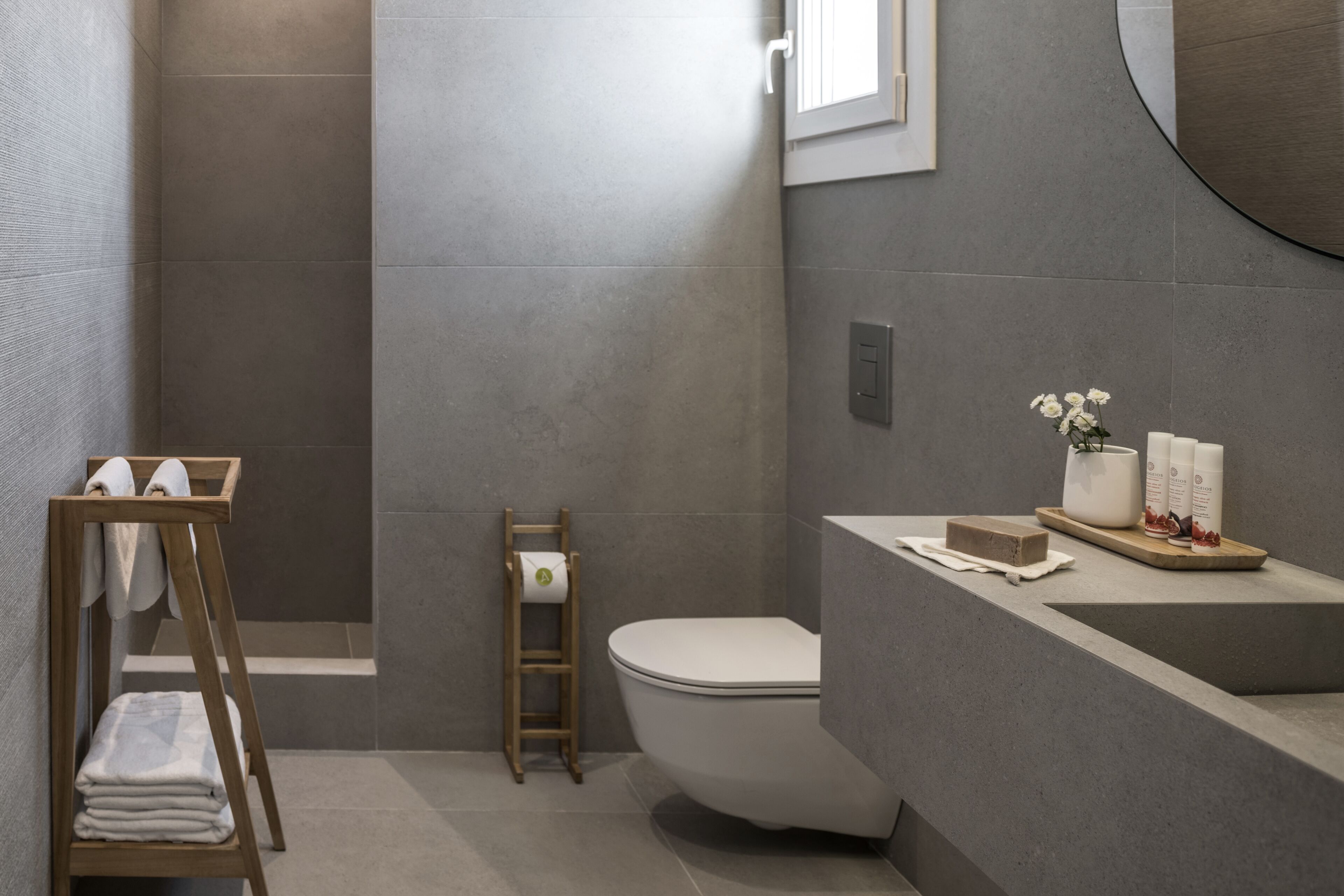 A modern bathroom with a toilet, a wooden towel rack, a shower area, and a vanity with toiletries.