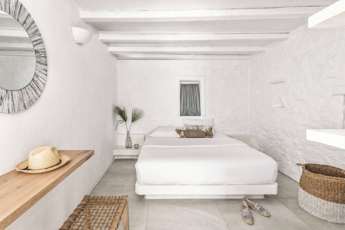 A bedroom with a bed, a bedside table, a mirror, a window, and a wall-mounted shelf.