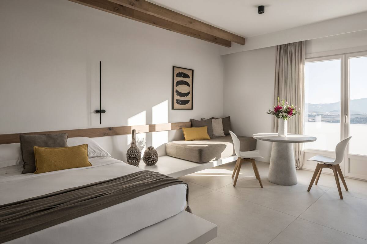 A modern hotel room with a large bed, a round dining table, and a view of the landscape through the window.