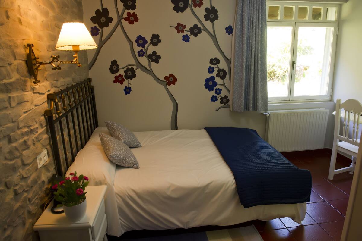 Un dormitorio con cama, mesita de noche con lámpara, un mural de pared con flores y una ventana con cortinas.